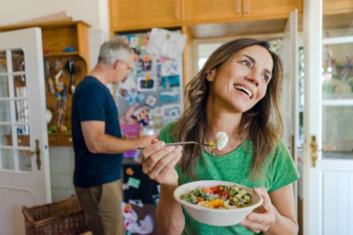 happy mature woman at home eating a salad with man in background - food stock pictures, royalty-free photos & images