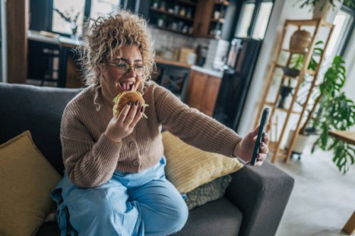 happy woman enjoying a hamburger on the couch while taking a selfie - junk food stock pictures, royalty-free photos & images