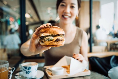 happy young asian woman sitting in a restaurant having her lunch. she is eating cheese burger with fries and a cup of coffee. enjoying a relaxing afternoon - junk food stock pictures, royalty-free photos & images