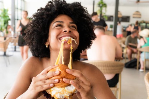 happy young woman enjoying a delicious burger at a restaurant during summer vacation - junk food stock pictures, royalty-free photos & images