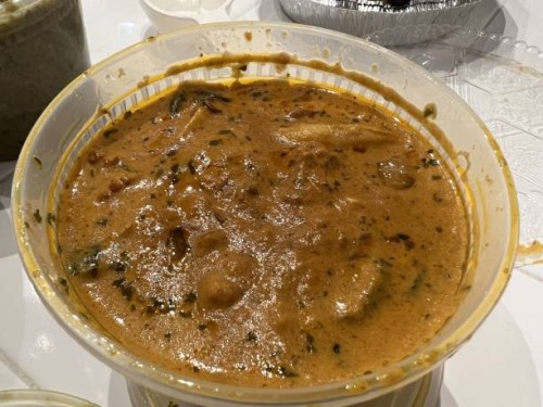 High-angle shot of a plastic carry-out bowl filled with meat and cooked green vegetables in a rich brown gravy, resting on a white tiled surface in...