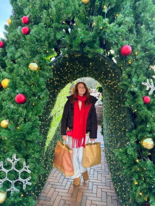 image of attractive, redheaded woman wearing black puffer jacket, aran jumper, red scarf and faux fur earmuffs, carrying brown paper shopping bags, walking on brick paved path under artificial conifer hedge arch, christmas ba
