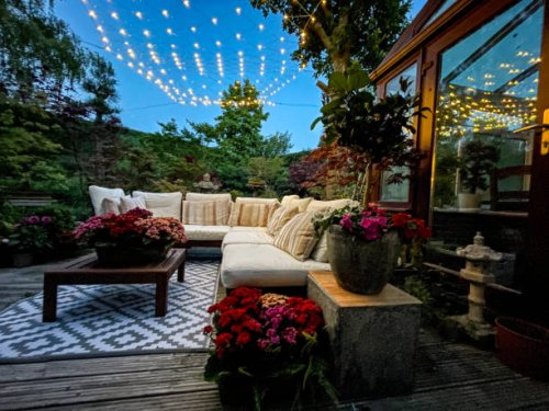 image of outdoor lounging area at night illuminated by string fairy lights, hardwood seating with cushions, wooden table top with flowering plant centrepiece, bonsai trees, japanese maples, landscaped oriental design garden, 