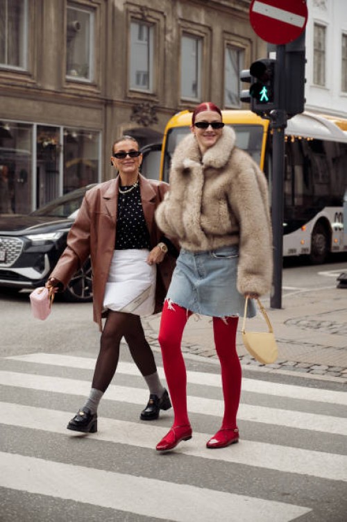 Janka Polliani wears black loafers, grey socks, a white Prada pillow skirt, a black top, and a brown leather jacket and Marianne Theodorsen wears red...