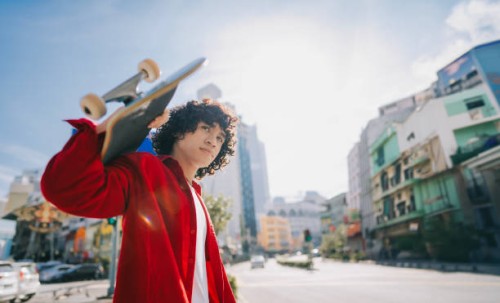 joven malayo asiático que lleva patineta cruzando la calle de la ciudad el fin de semana por la mañana - fashion fotografías e imágenes de stock