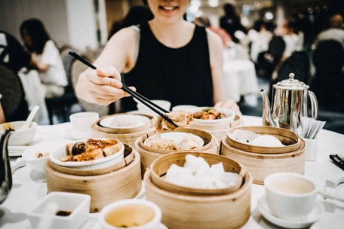 joyful asian woman picking up steamed beancurd roll with chopsticks and enjoying a variety of freshly made dim sum in restaurant - junk food stockfoto's en -beelden