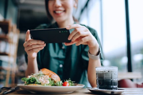 joyful young asian woman taking photos of delicious lobster salad brioche bun with bamboo charcoal black and white colour coffee with smartphone before eating it in cafe. eating out lifestyle. camera eats first culture - food