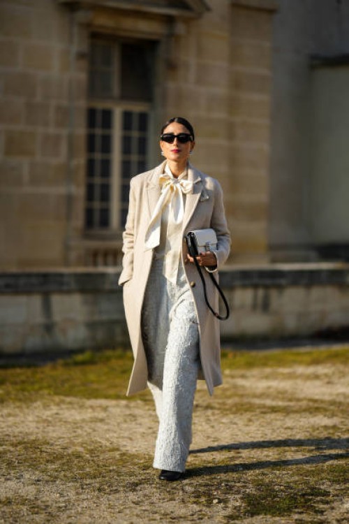 Julia Haghjoo wears black sunglasses, silver large pearl earrings, a white latte silk knot neck blouse, a beige long wool coat, a white embroidered...