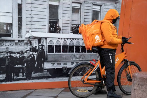 Just Eat takeaway delivery cyclist checks messages while standing next to a poster from the Victorian era, during the third lockdown of the...