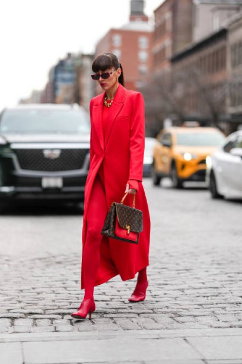 Katya Tolstova wears dark red sunglasses, gold hoop earrings, gold necklaces, bright cherry red blazer jacket, bright cherry red long sleeve sweater,...