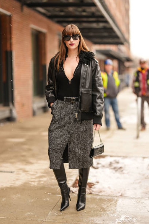 Koukla Lapidus wears black sunglasses, black half unzipped sweater, shiny black oversized leather jacket, dark gray pattern buttoned high waisted...