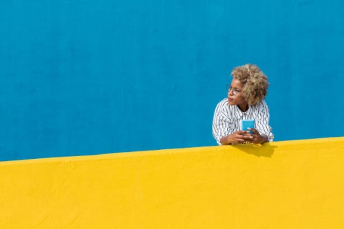 latin woman holding cell phone leaning on yellow wall over blue background. technology and social media concept. banner template, copy space for text on the side. - fashion fotografías e imágenes de stock