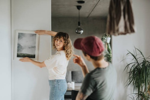 lesbian woman taking advice from girlfriend while hanging frame on wall at home - home decoration stock pictures, royalty-free photos & images