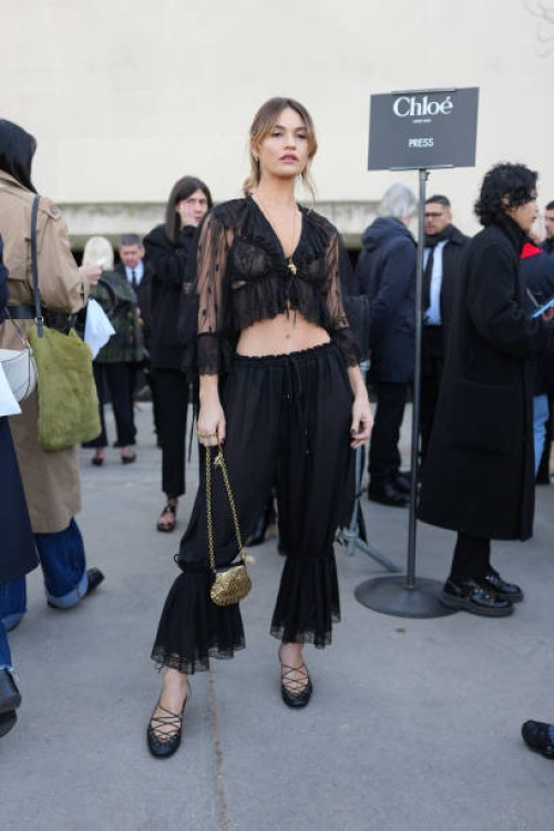 Lily James wears gold earrings, black sheer mesh cropped Chloe top, white black lace bra, gold necklace, black sheer mesh Chloe pants, gold shell...