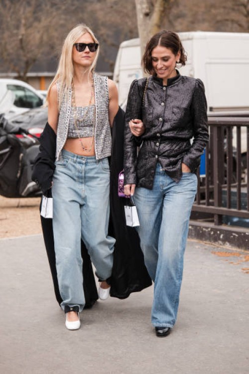 Linda Tol wears grey Chanel top with matching vest, blue jeans, black coat, white Chanel heels, and Erika Boldrin wears black sequins jacket, black...