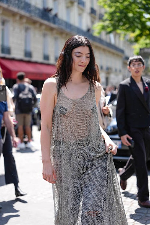 Lorde wears silver sheer mesh Balenciaga maxi dress, light blue baggy denim jean pants, shiny black pointed toe heels leather shoes, a silver ring,...