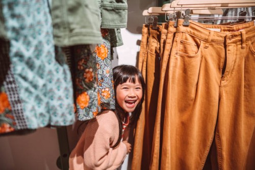 lovely little girl smiling joyfully behind a clothes rack while shopping in a clothing store - fashion stock pictures, royalty-free photos & images