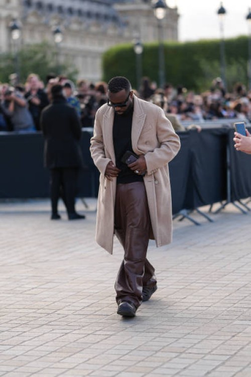 Maitre Gims wears black sunglasses, light brown blazer jacket, shiny dark brown leather pants, dark brown leather shoes, silver rings, outside Louis...
