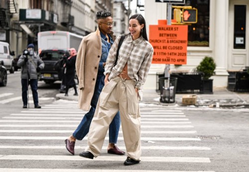 Mal Wright is seen waring a tan coat, jean top and pants and gold earrings with clear glasses with Rae Sada wearing a plaid shirt, tan pants and...
