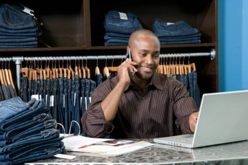 male employee behind desk in clothing boutique working on computer and talking on cell phone - home decoration stockfoto's en -beelden