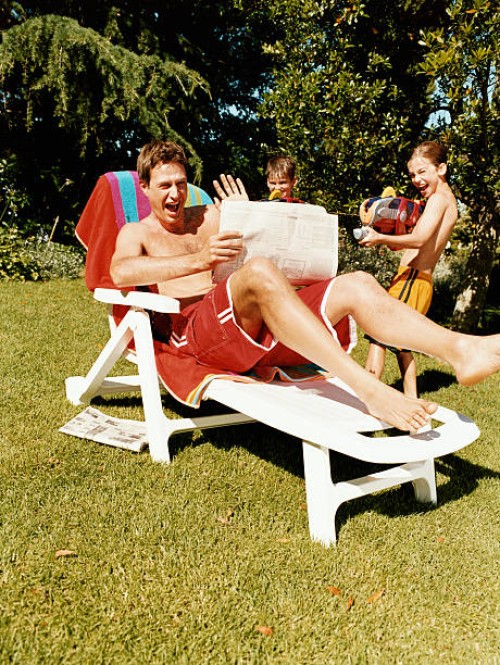man sitting on a sun lounger with a newspaper is surprised by two boys with water-guns - garden decoration photos et images de collection