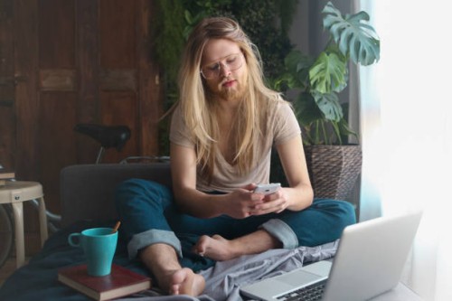 man with long hair and beard sitting on sofa bed looking at smartphone - fashion stock pictures, royalty-free photos & images