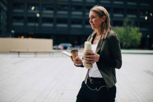 mature businesswoman carrying food and drink with laptop while walking on footpath in city - junk food stock pictures, royalty-free photos & images