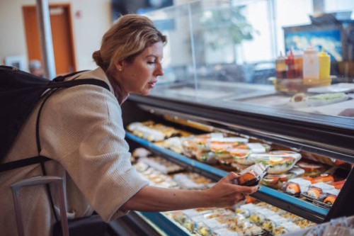 mature woman in casual clothes buying sushi at a supermarket, standing at a display of packaged food, focused on choosing, reflecting modern consumer lifestyle. - food stock pictures, royalty-free photos & images