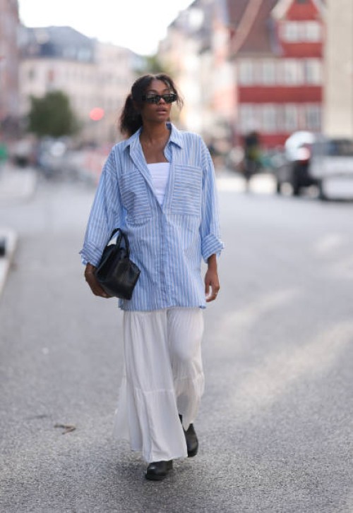 Maureen Adim is seen wearing black sunglasses with wide temples, hanging earrings with blue stone, an oversized blue and white striped shirt with...