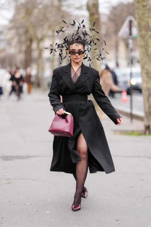Merve Gorgotz wears black feather head piece, black sunglasses, silver earrings, dark brown sheer mesh dress, black wrapped blazer coat, shiny...