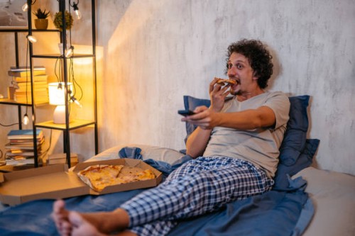 mid-adult man eating pizza and watching tv in the bed at night - junk food stock pictures, royalty-free photos & images