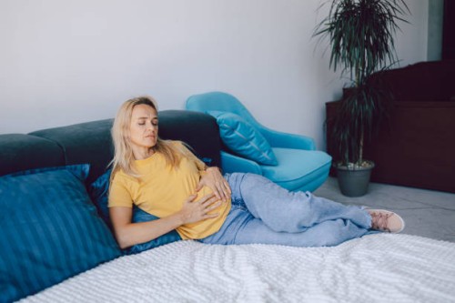 middle-aged woman holds her hands on her stomach while lying on a bed at home. pain, negative emotions and suffering. - junk food stock pictures, royalty-free photos & images