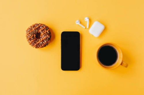 minimalistic breakfast scene with coffee, a chocolate donut, and smartphone on a bright yellow background - junk food stock pictures, royalty-free photos & images