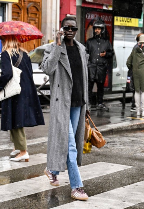 Model is seen wearing a gray coat, black sweater, light blue jeans and Adidas sneakers with brown bag and black sunglasses outside the Mugler show...