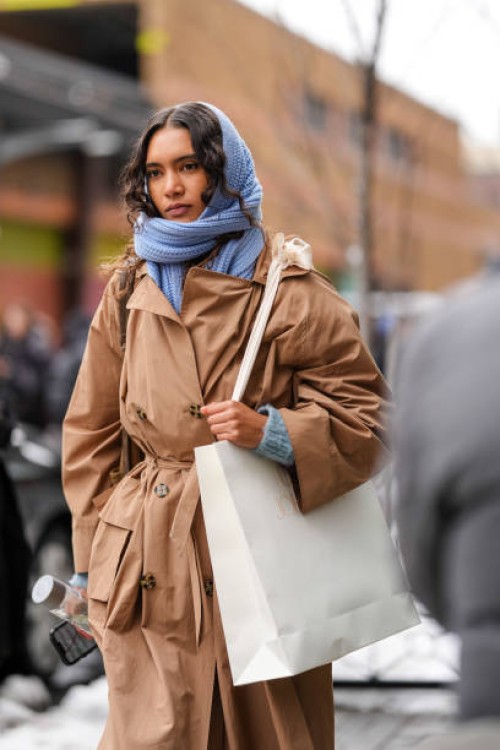 Model wears a blue knit scarf over the head as a hood / balaclava, a brown trench coat, holds a paper shopping bag outside Ulla Johnson , during New...