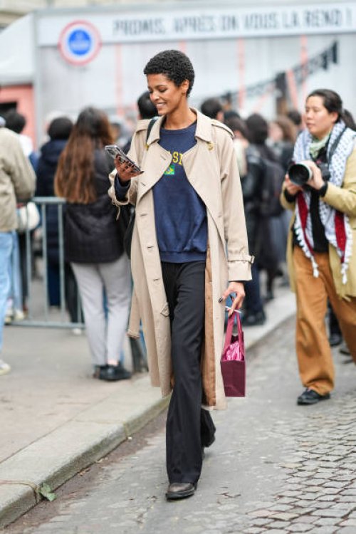 Model wears a dark blue pullover, a beige trench coat, black pants, brown and black leather shoes, a shopping bag, leather shoes, outside Nina Ricci,...