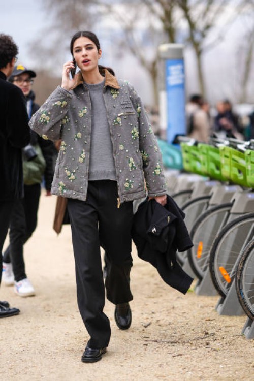 Model wears a grey pull-over, a grey jacket with yellow floral details, wide-leg black pants, leather shoes, outside Chanel, during the Womenswear...