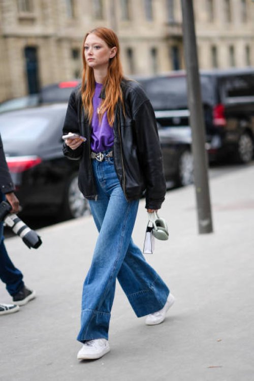 Model wears a purple t-shirt , a black leather jacket , a belt, blue flared denim pants / jeans , white sneakers shoes , outside Chanel, during the...