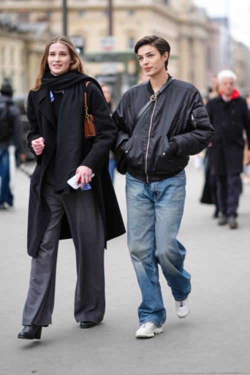 Model wears a scarf, a black coat, gray flared pants ; a model wears a black bomber jacket, blue denim pants / jeans , white sneakers shoes, outside...