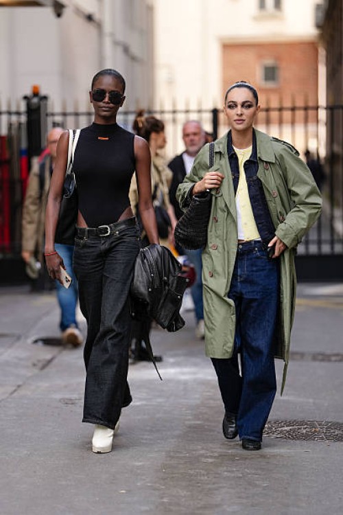 Model wears a sleeveless black top with a high neckline, paired with high-waisted dark denim jeans. Accessories include large black sunglasses and a...