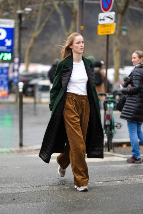 Model wears a white t-shirt, a green long coat with black quilted lapels, brown flared corduroy pants, sneakers shoes, outside Chanel, during the...
