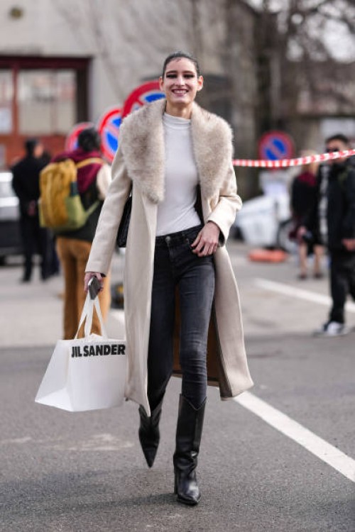 Model wears black eye shadow, white long sleeve shirt, cream wool coat with light gray faux fur collar, a black denim jean skinny pants, shiny black...