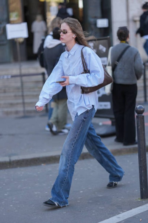Model wears black sunglasses, light blue buttoned up long sleeve shirt, shiny dark brown leather bag, navy blue washed loose denim jean pants, black...