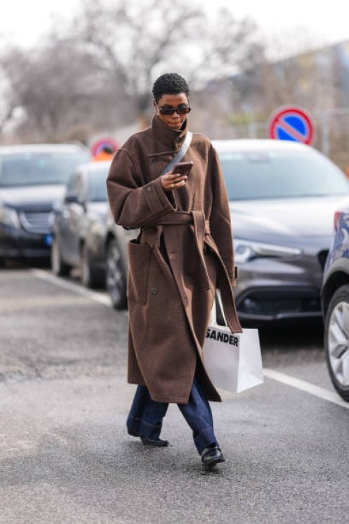 Model wears black sunglasses, silver earrings, dark brown wool oversized coat, a gold ring, black crossbody leather bag, dark navy blue lose denim...