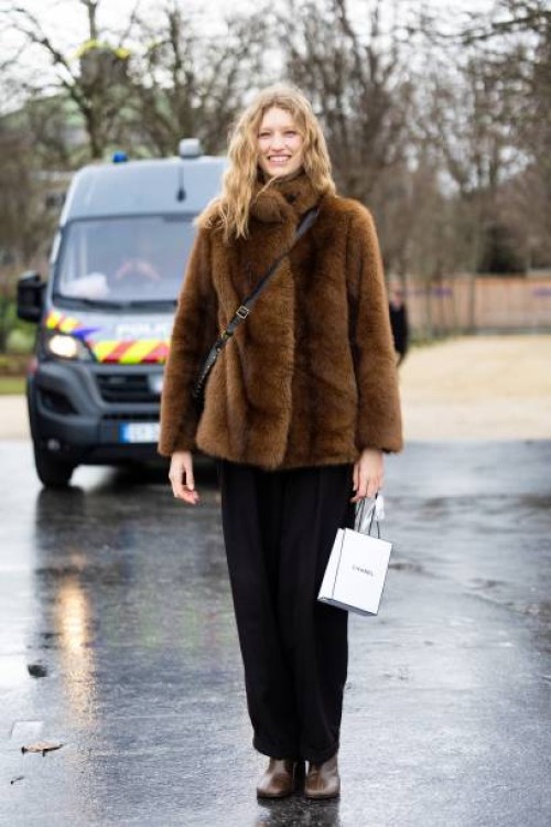 Model wears black trousers, brown heeled leather ankle boots, a black leather cross-body bag and a brown fur jacket outside Chanel during Haute...
