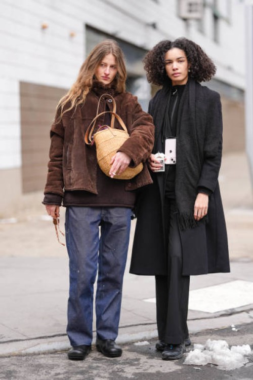 Model wears dark brown sunglasses, dark brown suede jacket, dark brown sweater, navy blue relaxed trouser pants, light brown woven basket, shiny...