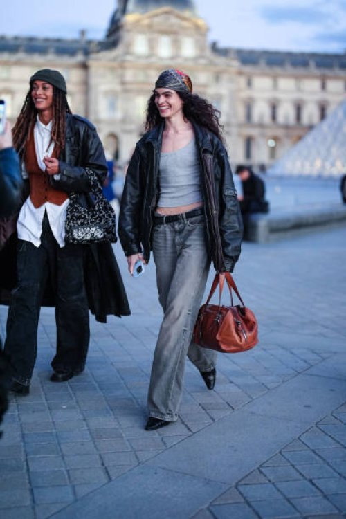 Model wears dark gray pattern head scarf, day gray tank top, shiny black oversized leather jacket, black leather belt, washed dark gray denim jean...