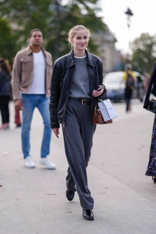 Model wears dark gray sweater, white t-shirt, shiny black oversized leather jacket, black leather belt, dark gray loose pants, shiny black loafer...