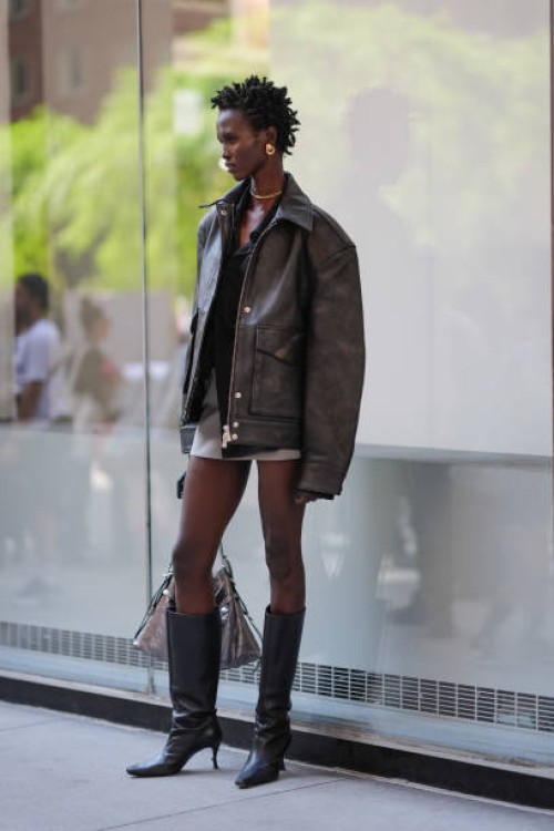 Model wears gold earrings, gold necklace, black shirt, dark brown oversized blazer jacket in leather, light gray mini skirt, shiny black pointed toe...