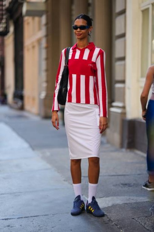 Model wears sunglasses, a red and white striped polo shirt, a white on-knee skirt, white socks, Adidas blue and yellow striped sneakers shoes,...